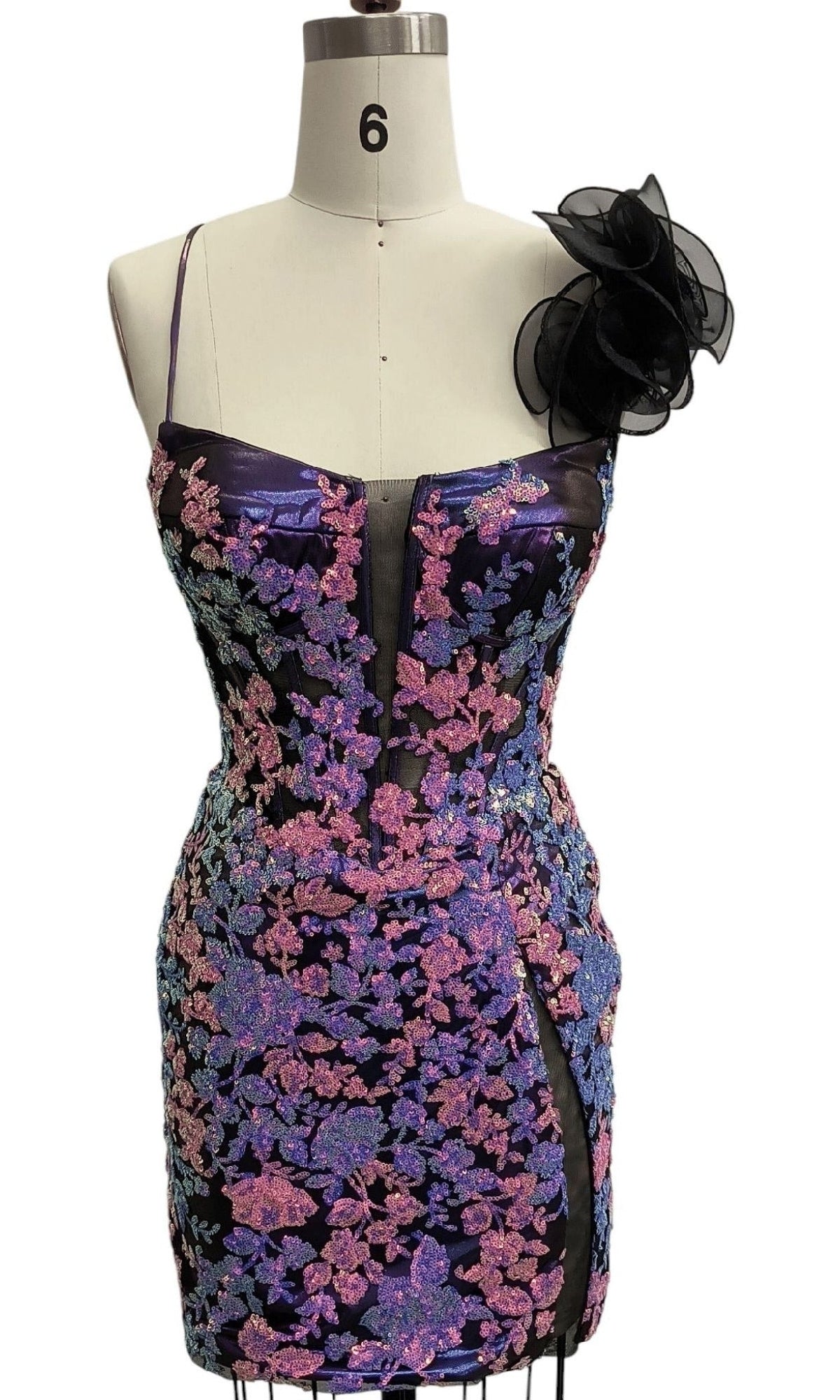 A mannequin displays the Short Purple Sequin Hoco Dress 12567, a dazzling strapless mini with sequin details, a sheer central panel, side slit, and an eye-catching floral accent on one shoulder.